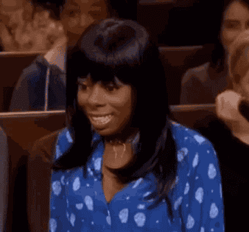 A woman in a patterned blouse smiles and looks surprised while sitting among a crowd