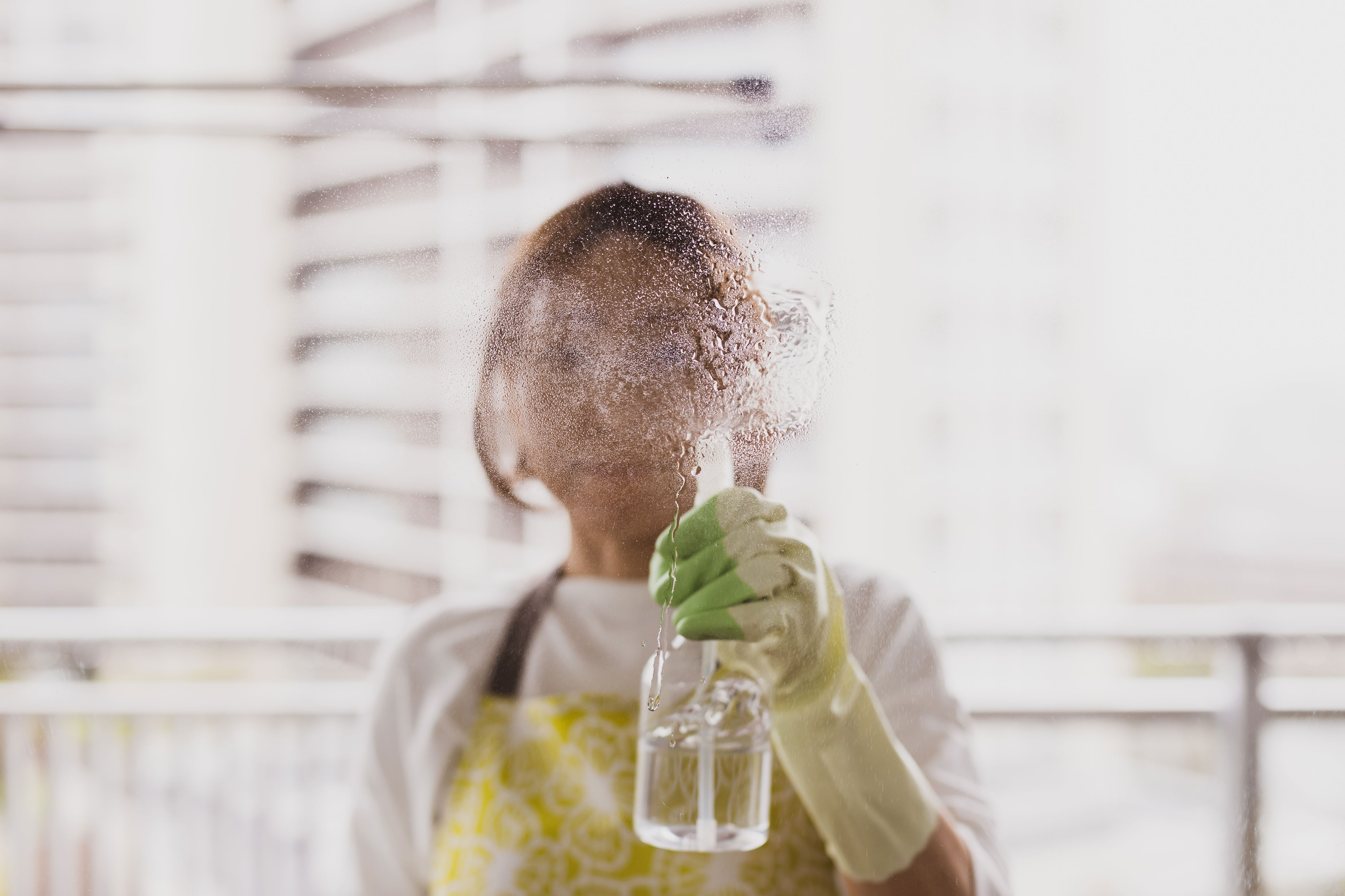 Person spraying water from a bottle on their face, wearing gloves and an apron, with a blurred cityscape in the background