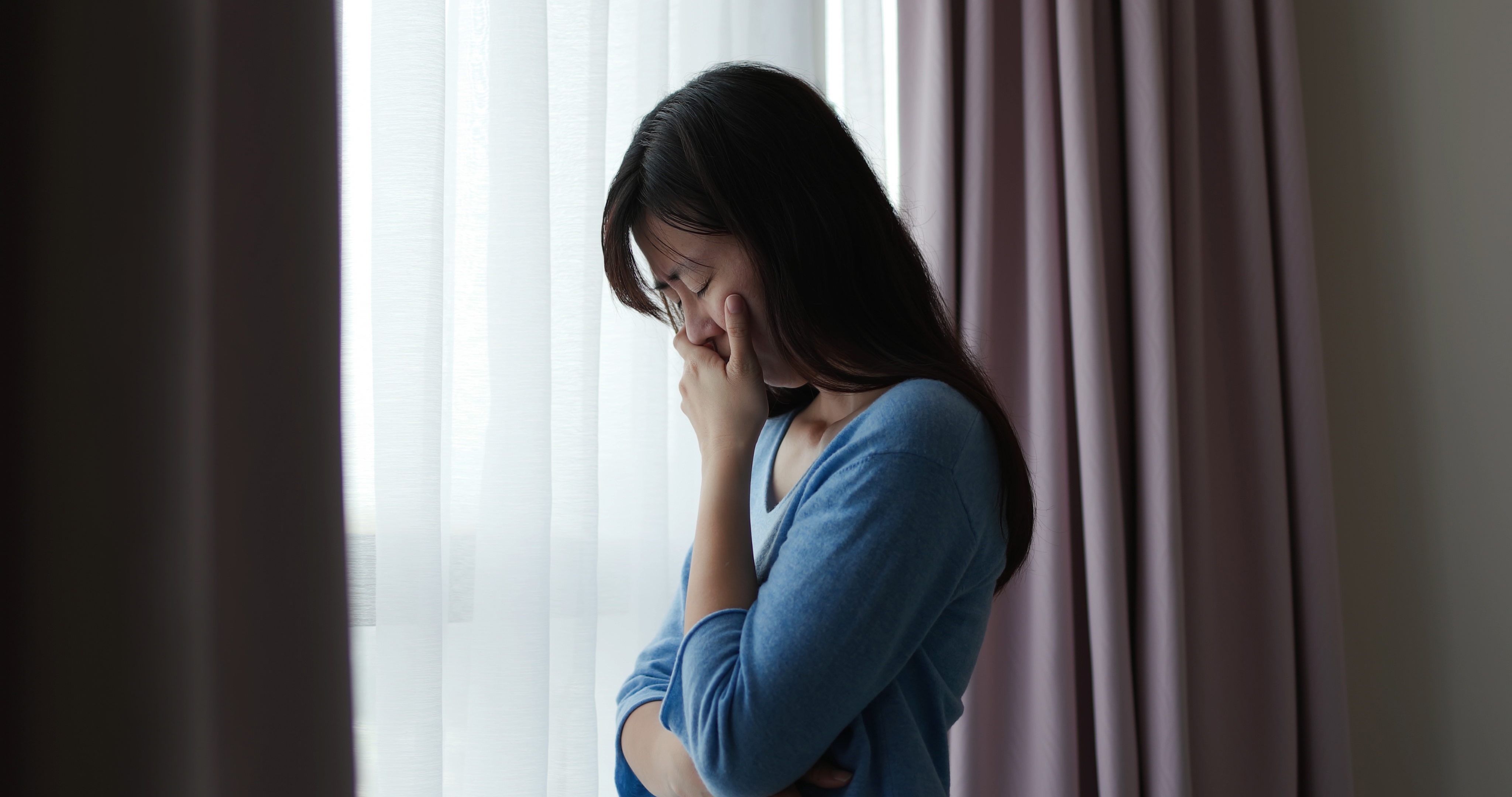 Person standing by a window with curtains, appearing thoughtful or concerned, hand near their face, wearing a comfortable long-sleeved top