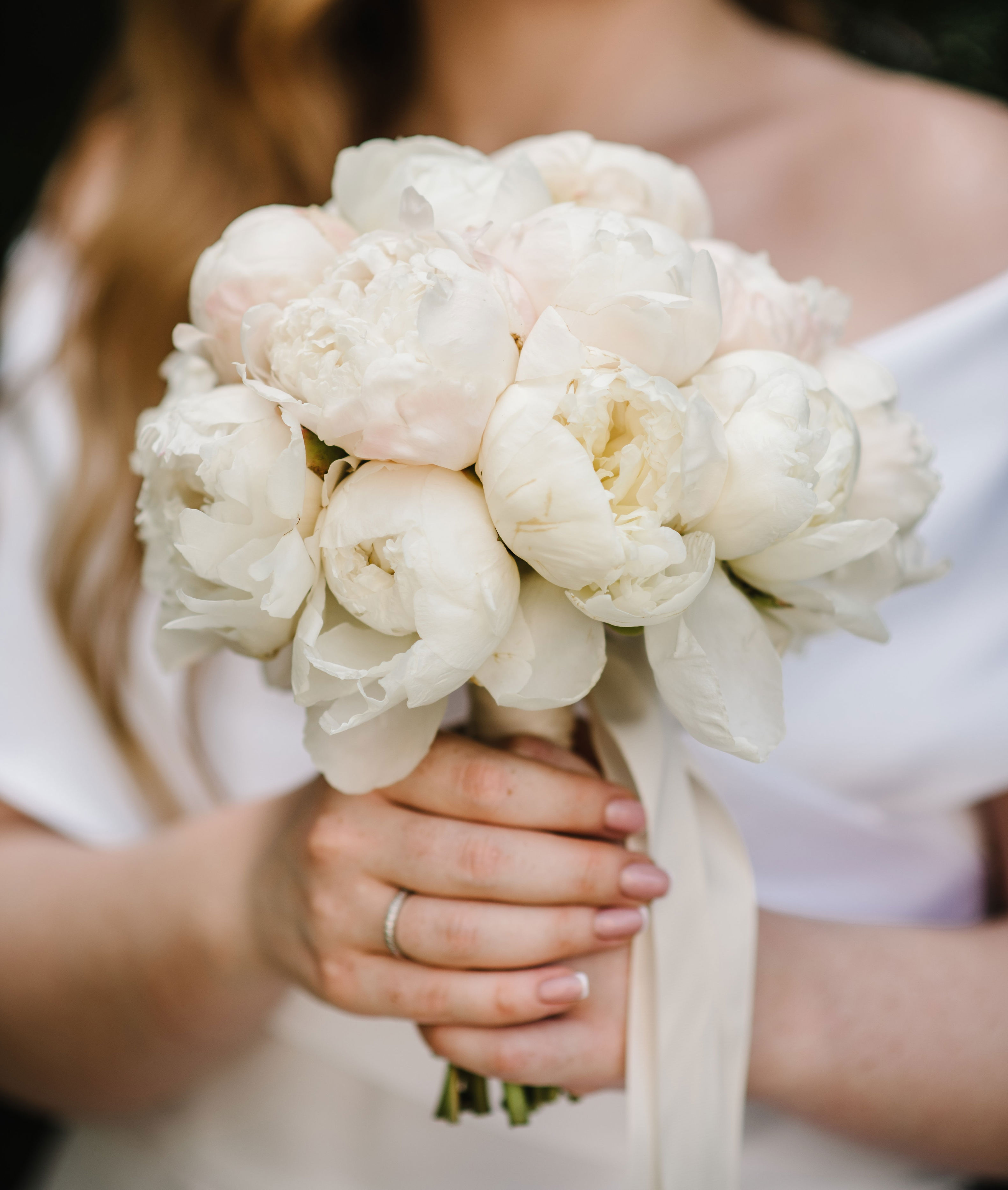 A bride holds a bouquet of white peonies, wearing an off-shoulder dress with loose hair, captured from shoulders down