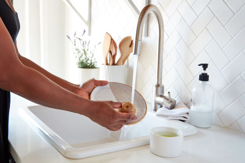 Person washing a plate under a kitchen faucet with soap dispenser and wooden utensils nearby.
