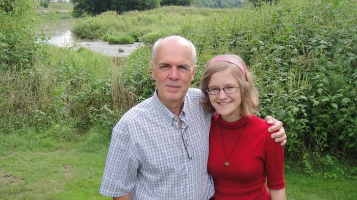 A man and a woman are standing in a grassy area next to a river. The man is wearing a plaid shirt, and the woman is wearing a dress with a headband