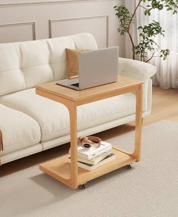 Wooden side table with wheels, holding a laptop, books, and headphones, positioned beside a sofa in a stylish living room setting