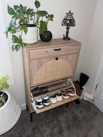 Wooden shoe cabinet with a woven front panel, topped with plants and a small stained glass lamp, displaying neatly organized shoes below