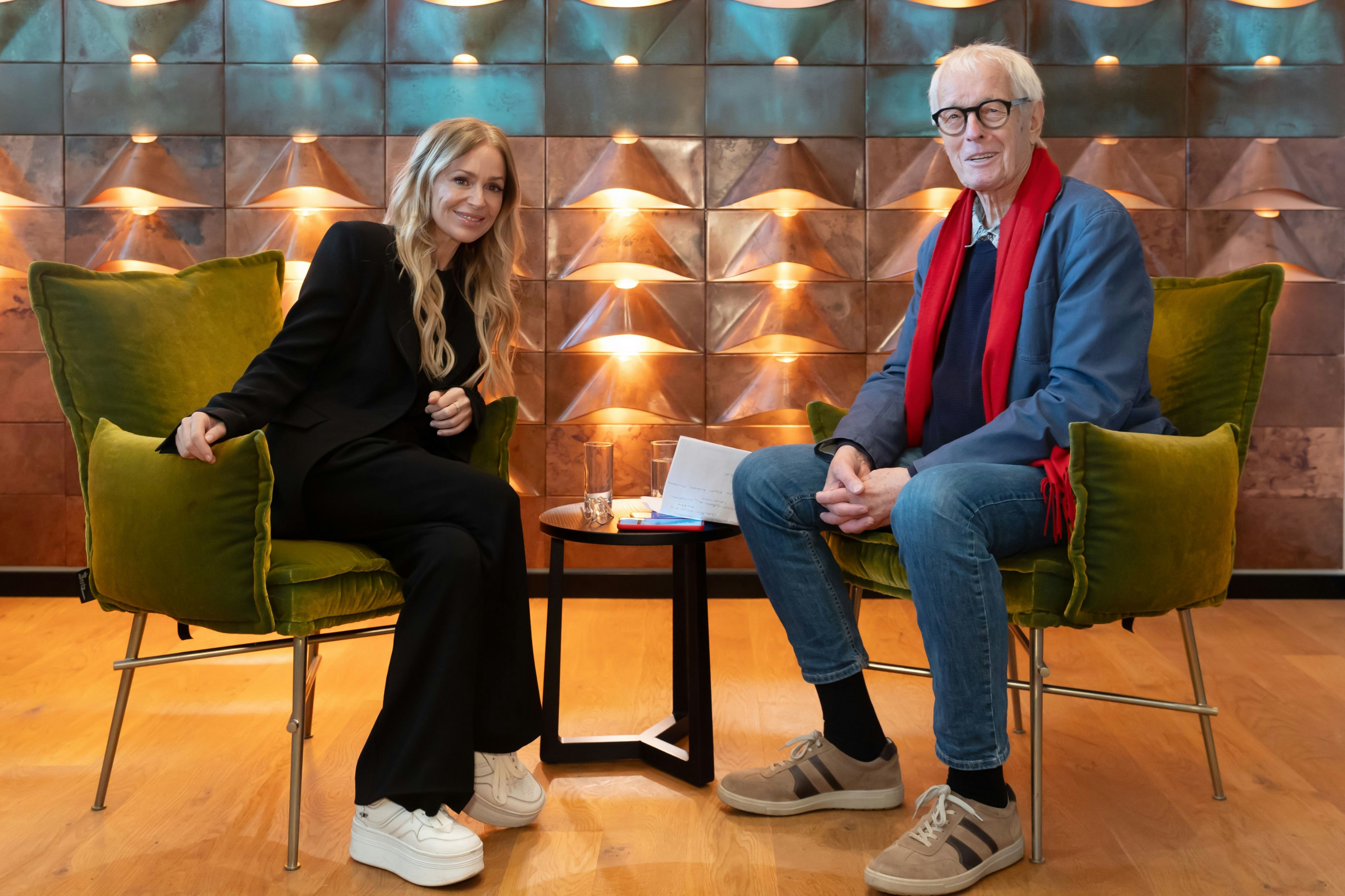 Eine junge Frau mit langen blonden Haare in einem schwarzen Hosenanzug sitzt einem älteren Herrn mit blauem Sakko und rotem Schal gegenüber. Beide sitzen vor einer golden angestrahlten Wand und lächeln.
