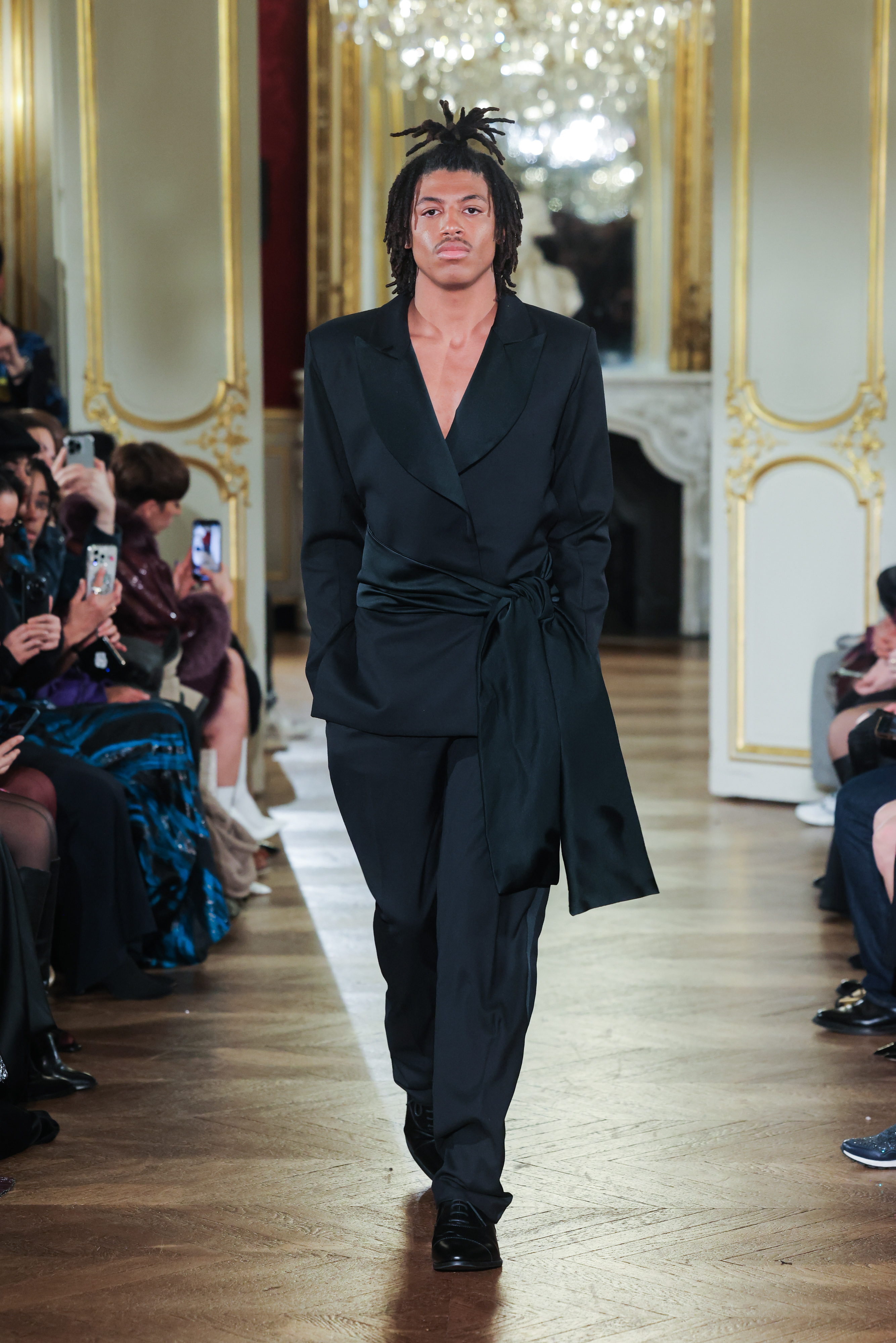 Henry Samuel walking the runway in a black suit with a large satin sash during the Lena Erziak Haute Couture Spring-Summer 2025 show.