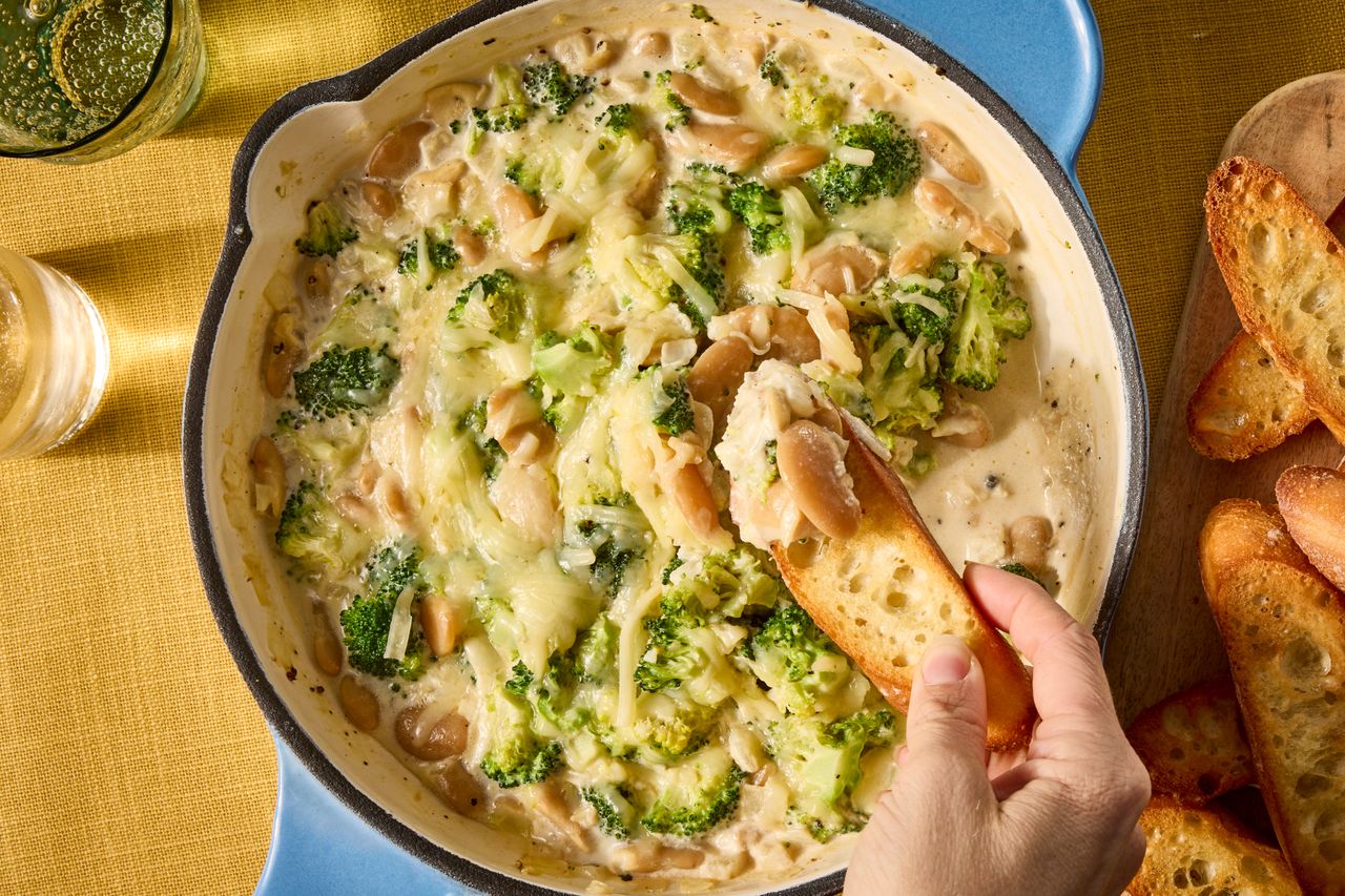 A skillet containing a creamy broccoli and bean dip being dipped into with a slice of bread by a hand