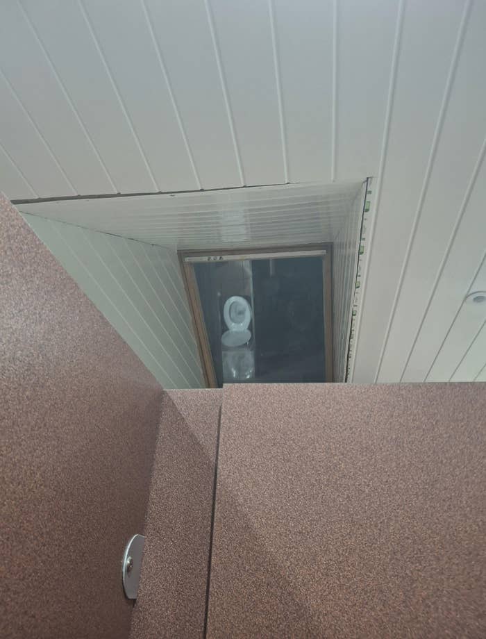 A bathroom ceiling panel is open, revealing a toilet with a roll of toilet paper on the seat