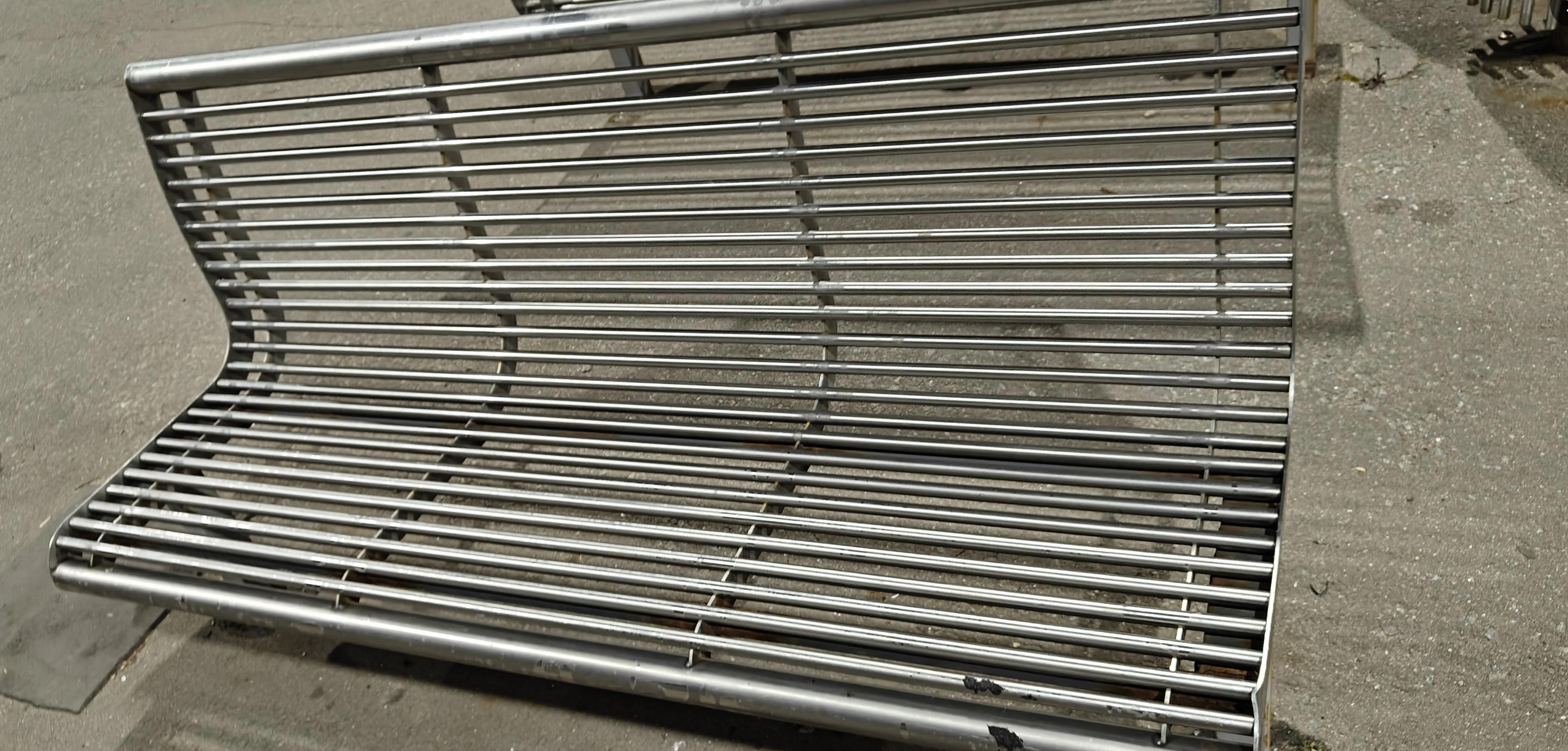 A metal bench with a modern, minimalist design, featuring horizontal bars set within a frame, is situated outdoors on a concrete surface