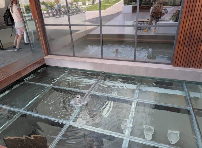 A small pool with a fountain under a glass walkway; in the background, a reflection of people and bikes is visible through the glass walls