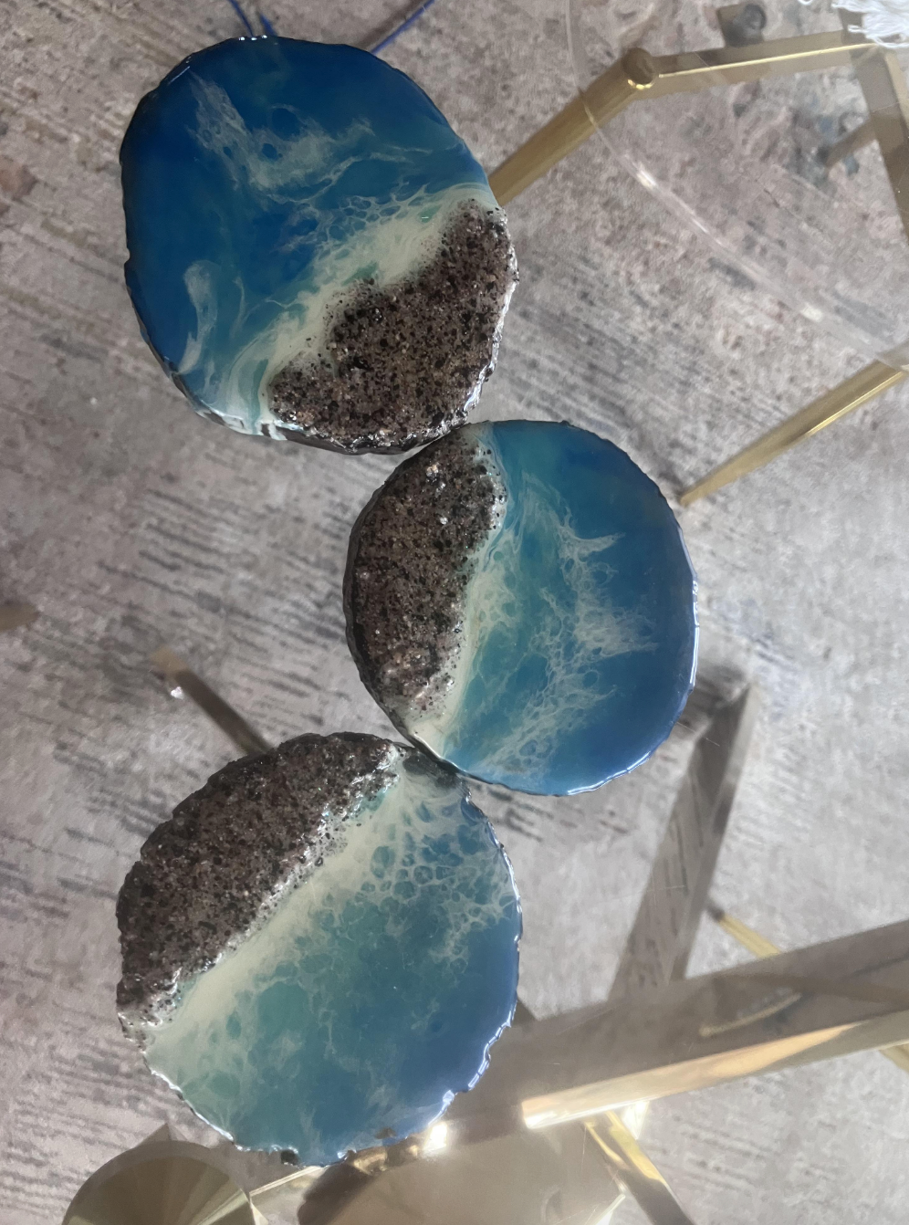 Three circular resin coasters with ocean-like designs, featuring blue and white waves next to a textured gray and brown section