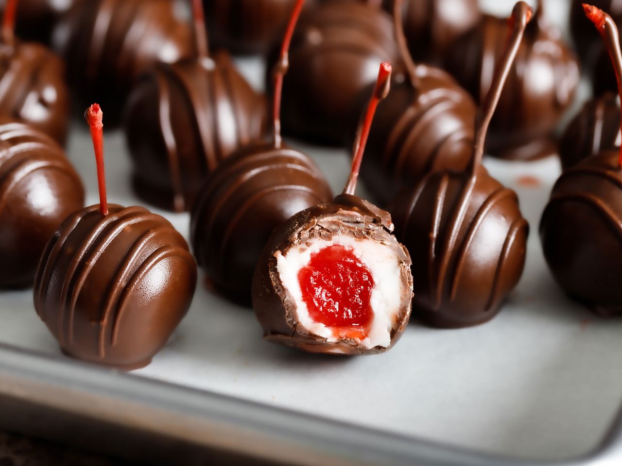 a close up view of several chocolate covered cherries chilling on a parchment lined baking sheet with one cut open revealing the creamy center wrapped around a bright red cherry.