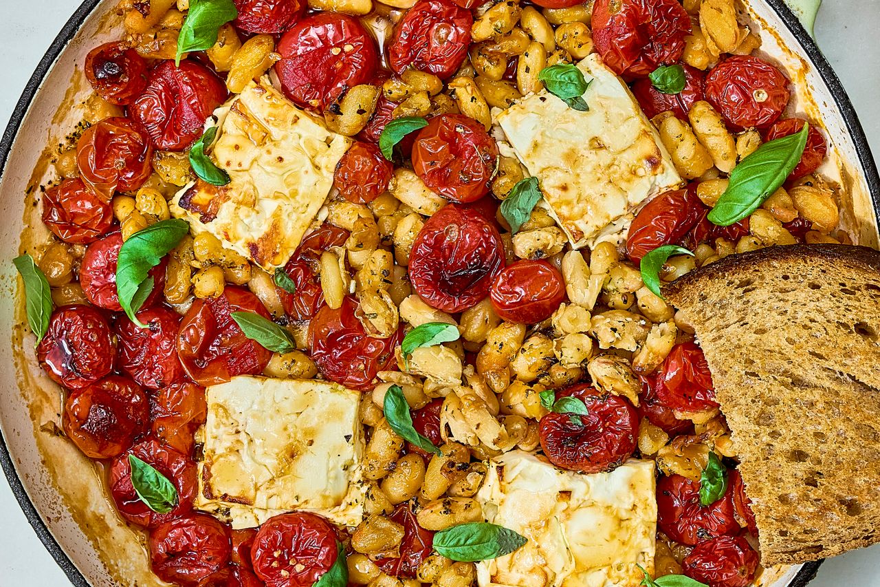 A skillet filled with baked feta cherry tomatoes white beans fresh basil leaves and a slice of toasted bread