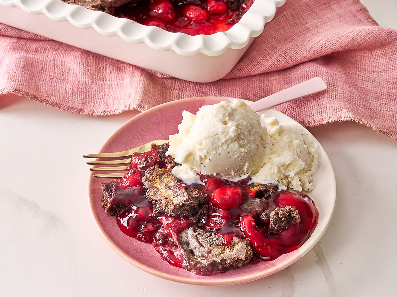 pink bowl of chocolate cherry dump cake with ice cream and with 9x13 pan in background on pink towel