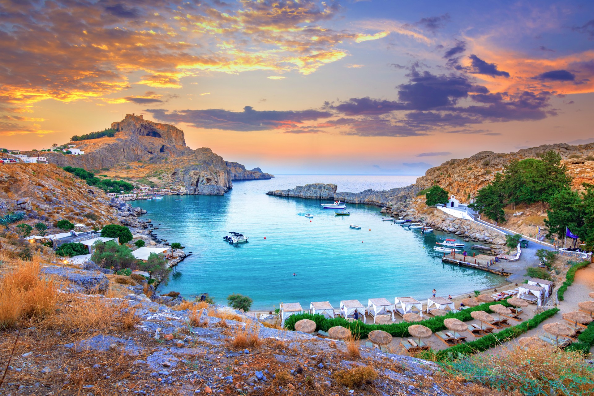 Panoramic view of a cove in Greece at sunset, surrouded by a rocky landscape. There ae boats on the water and sunloungers on the shore line.