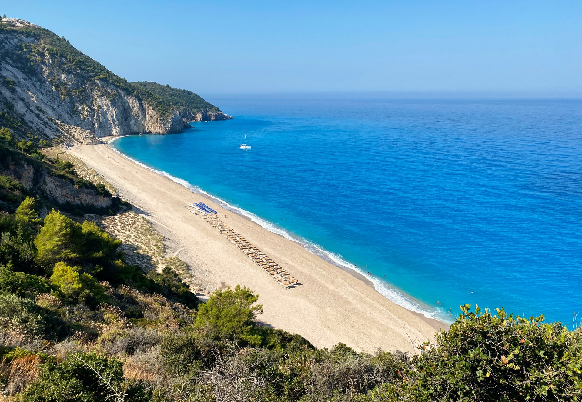 Aerial view of a large sandy beach with bright blue water and a row of sunloungers surrounded by greenery and under a blue sky.