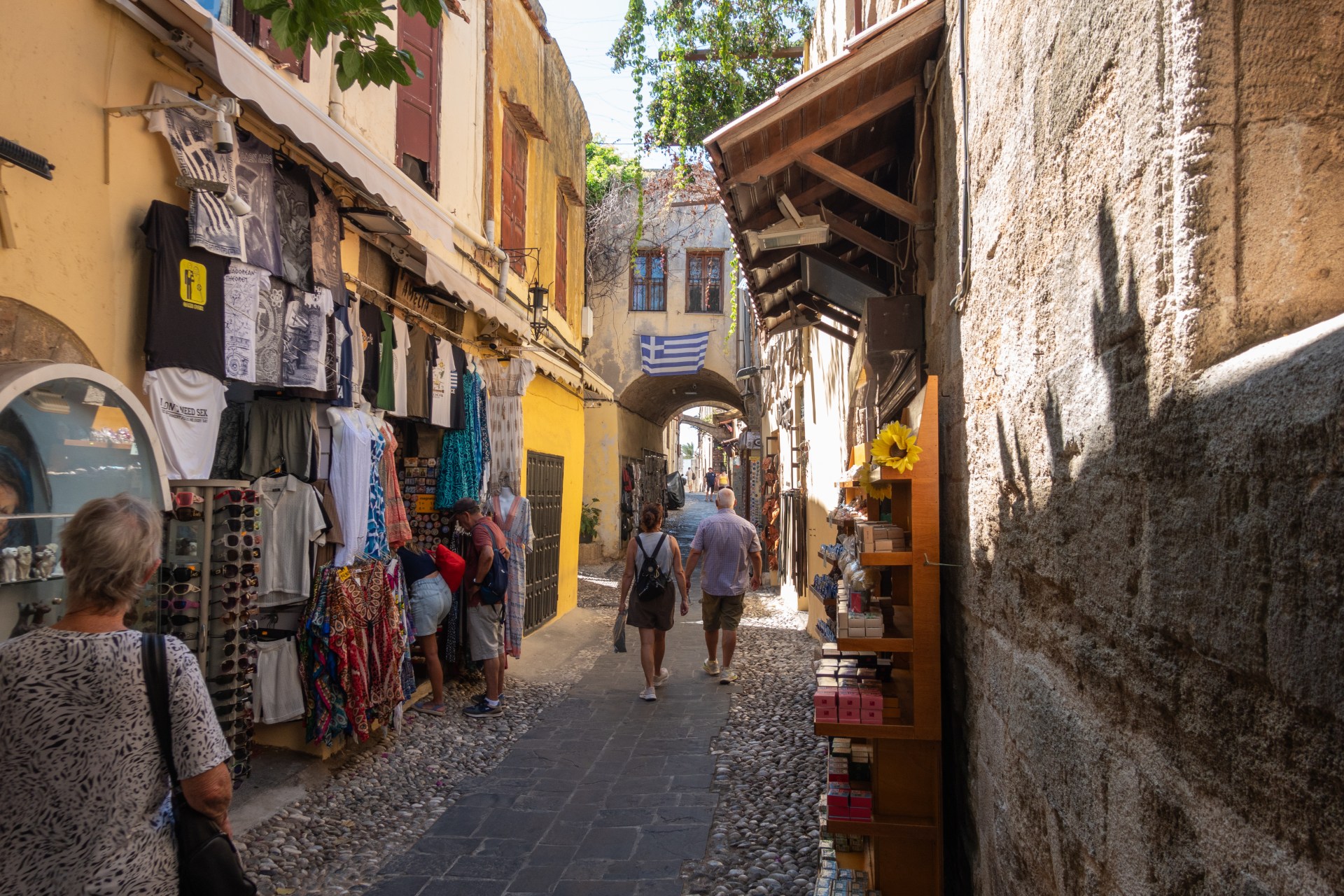 A narrow cobbled street lined with gift shops and an archway with a Greek flag above it.