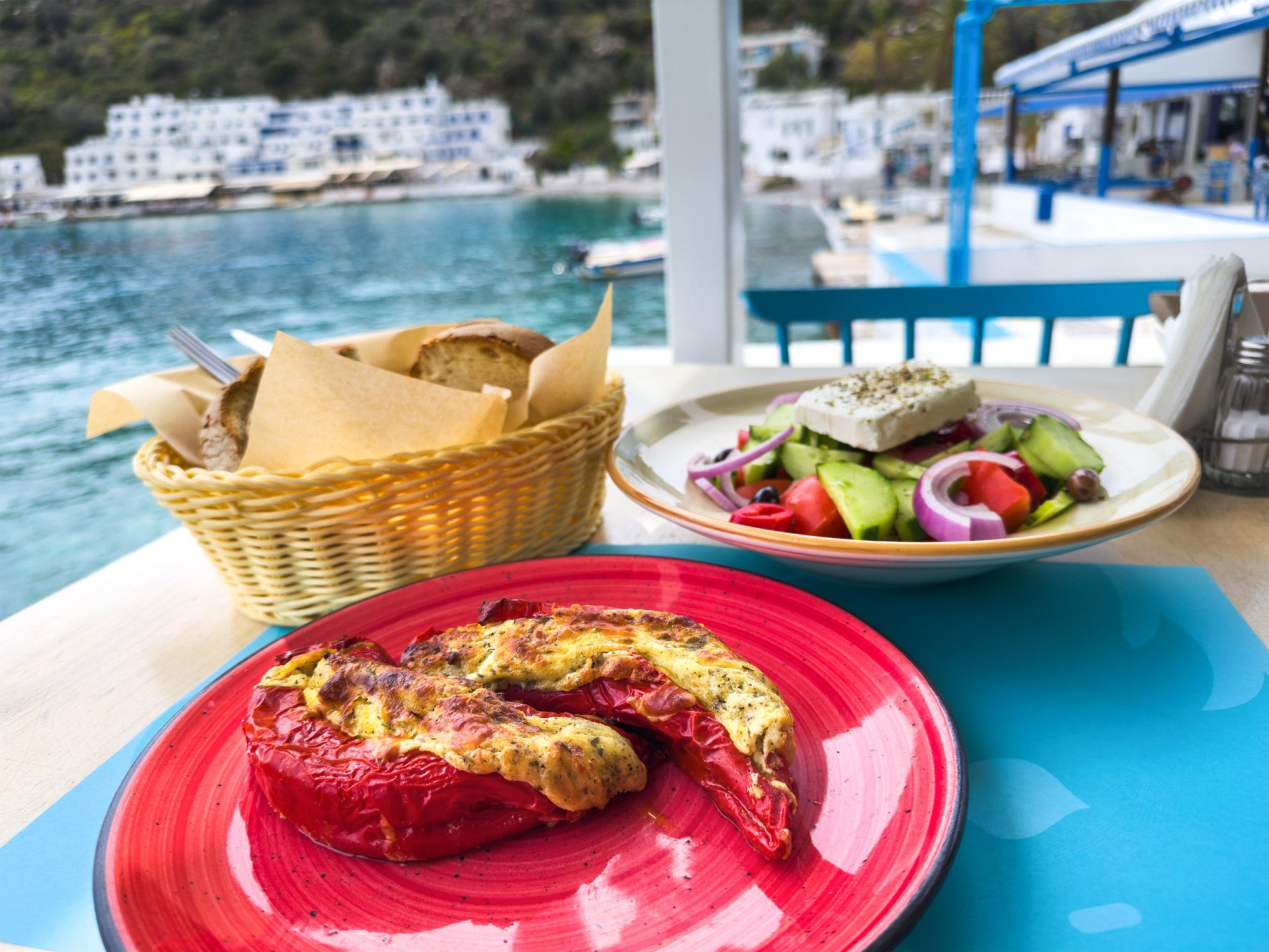Peppers filled with cheese, Greek salad and a basket of bread on a table wih a marina visible in the background.