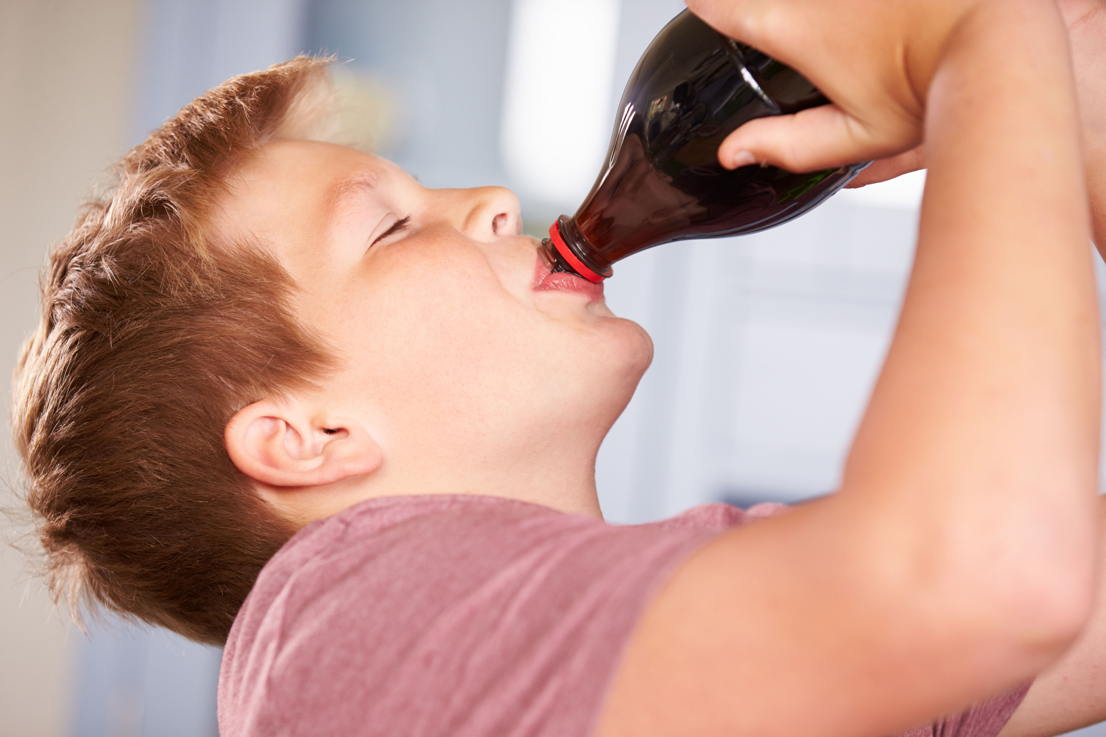 Child enjoys drinking from a large bottle, tilting head back. Relaxed, casual setting highlights playful interaction with a beverage