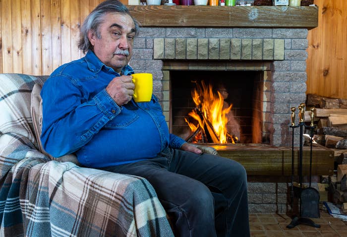 Older man in denim shirt holds yellow mug, seated by a fireplace on a plaid-covered chair, with logs stacked nearby