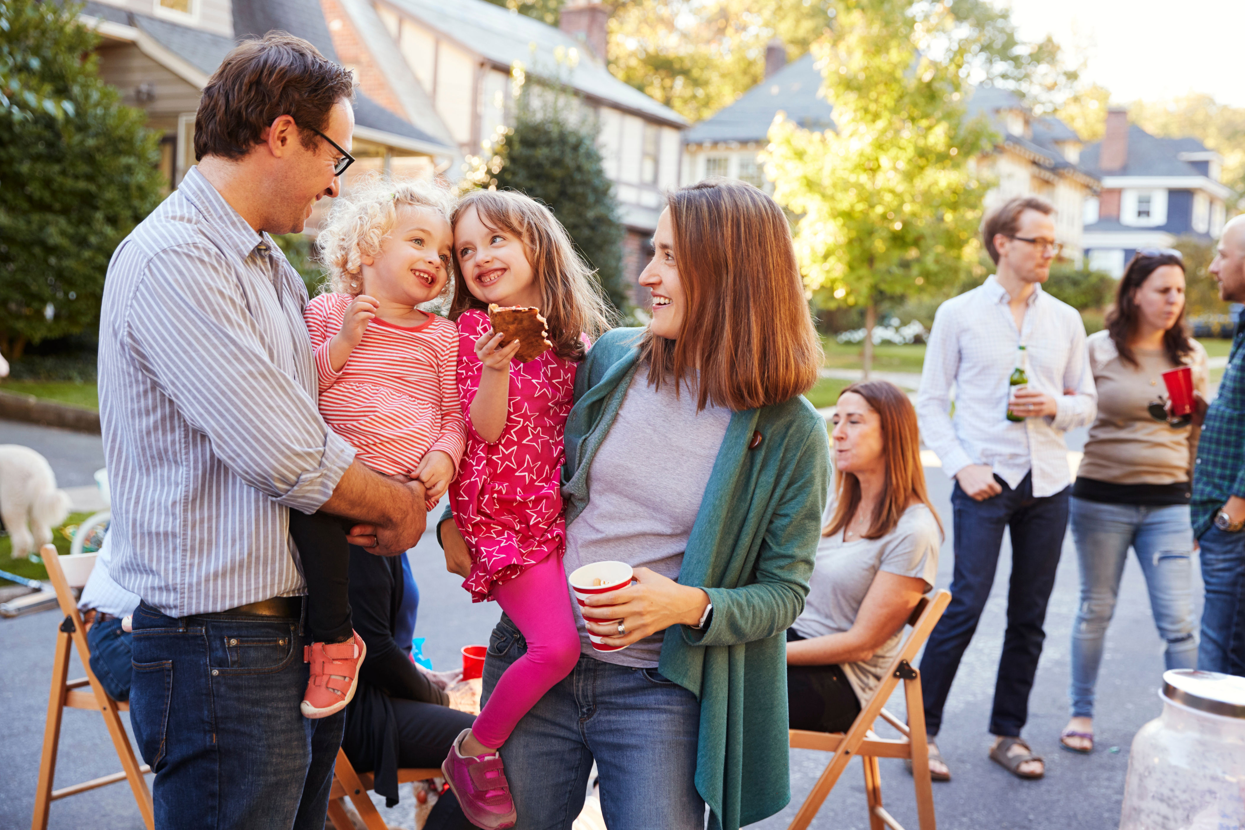 People at an outdoor gathering, two adults happily interacting with children. Others are seated or standing, holding drinks, in a neighborhood setting