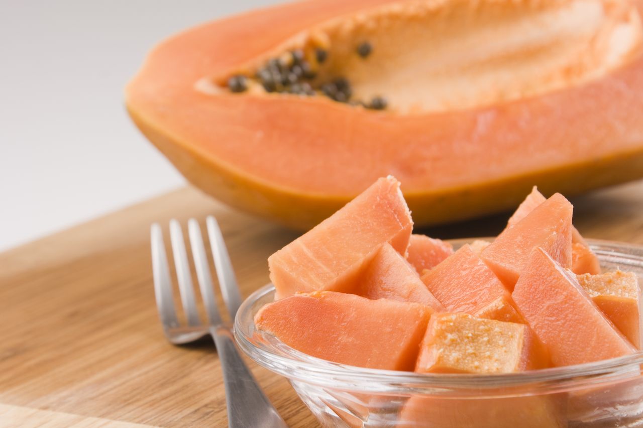 Close-up in focus shot of a glass bowl of papaya sliced in chunks, with a peeled whole fruit in the background.