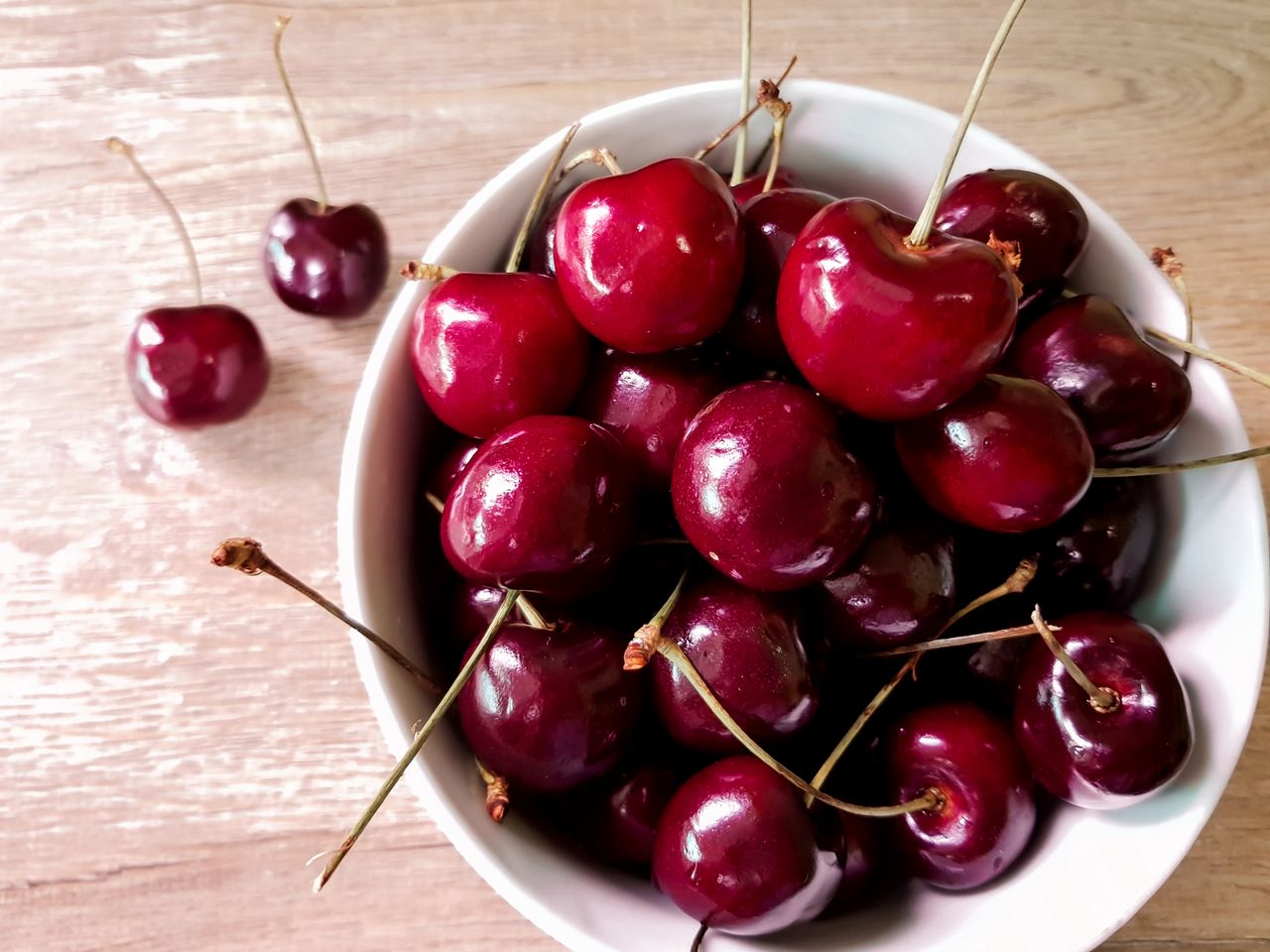 Top-down view of whole fresh cherries in a white bowl on a light wood table.