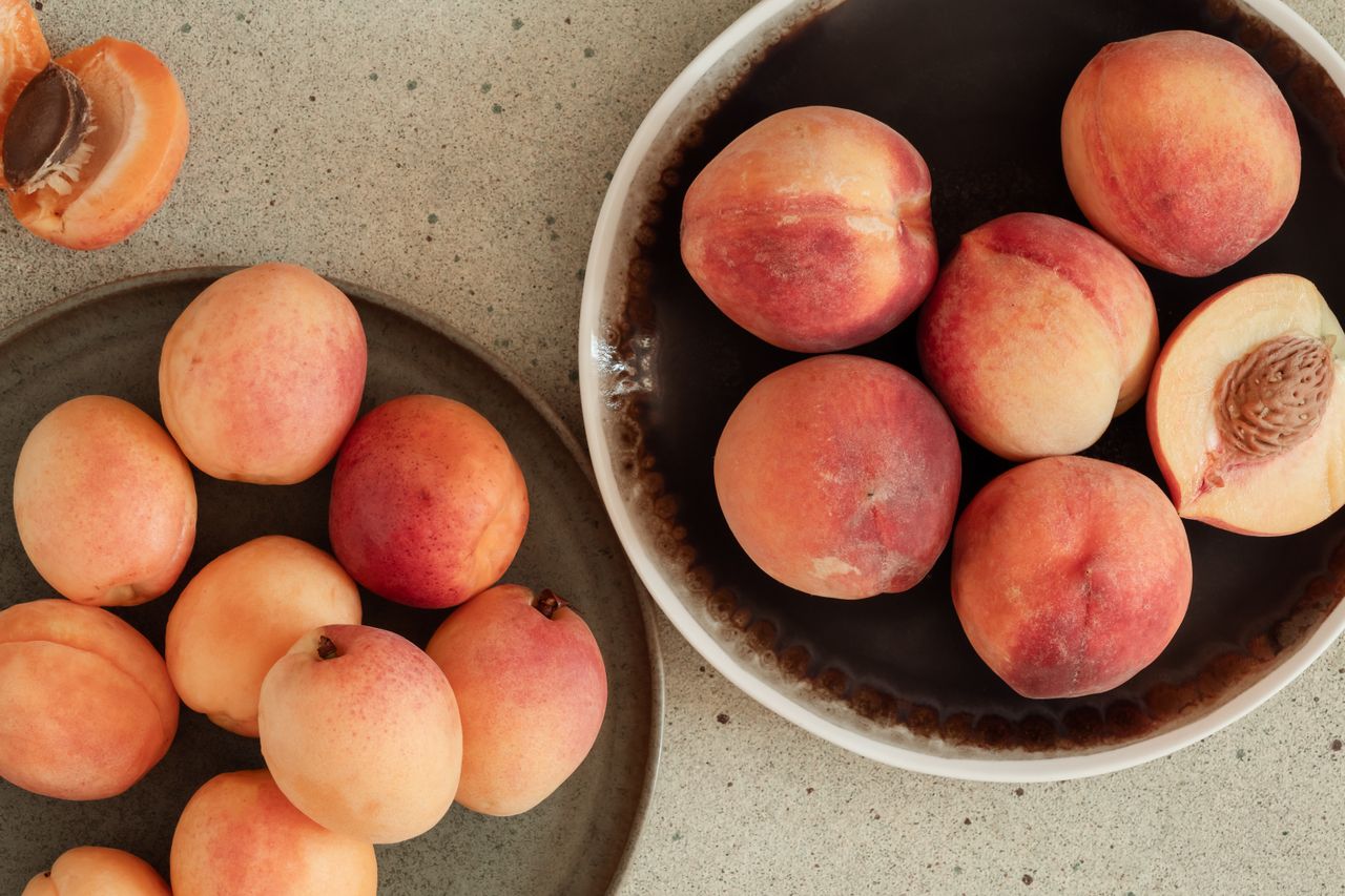 A bowl of fresh sliced and whole apricots with more whole apricots on a wooden plate next to it, arranged on a stone table.