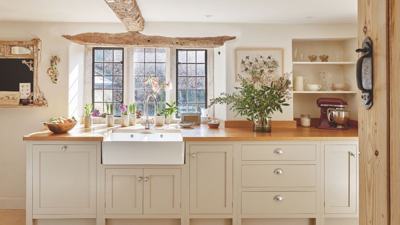A bright and open country-style cream kitchen with a white farmhouse sink set into a wooden counter below a bright window. Gray painted cabinetry with silver hardware is visible, as are original wooden beams on the ceiling and above the crittal window.