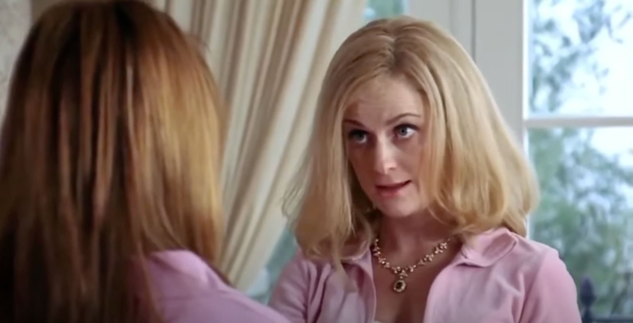 Two women are facing each other indoors. The woman facing us has short, styled hair and wears a necklace, appearing to be in a conversation
