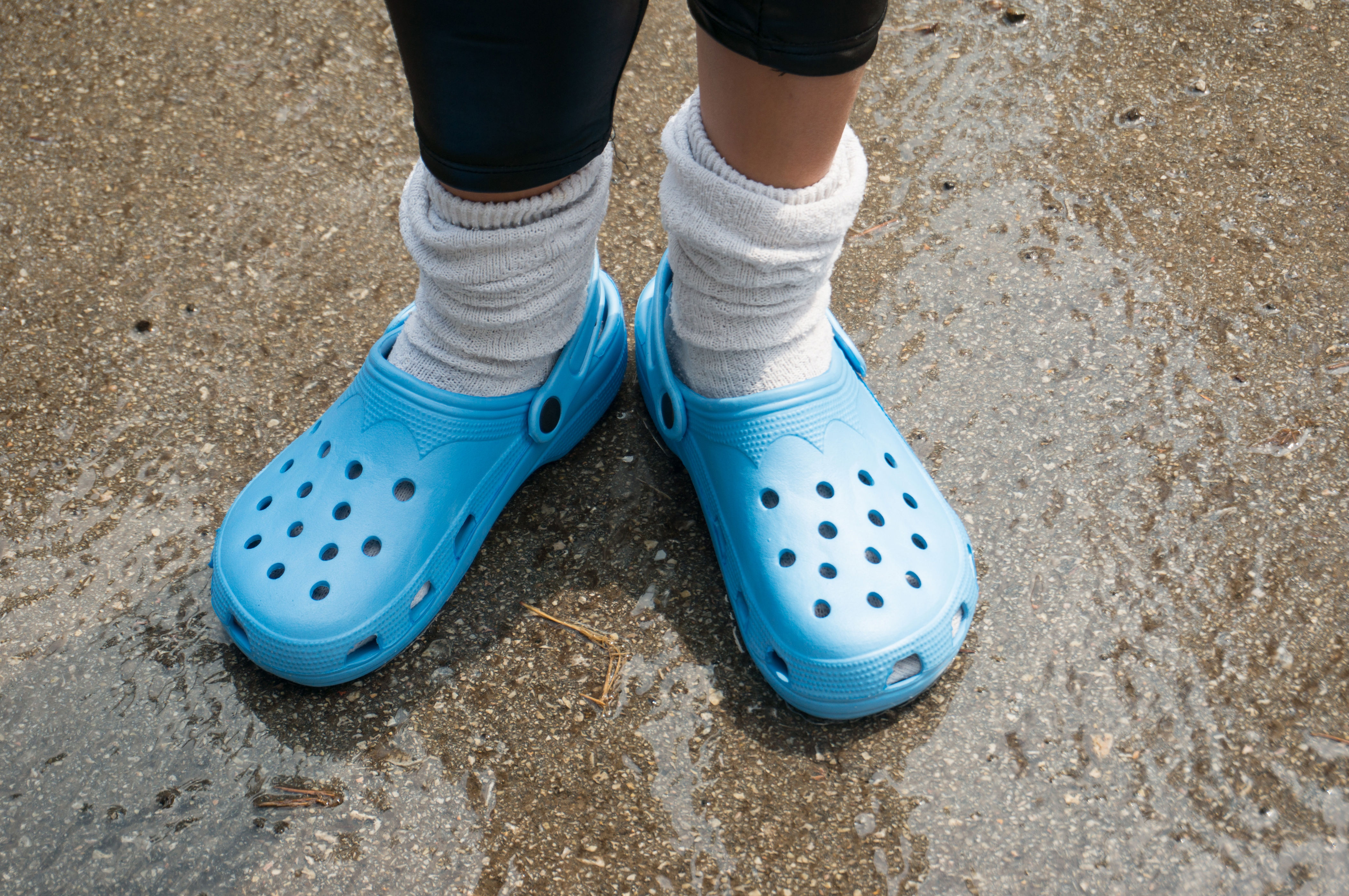 Person wearing white socks with blue clogs standing on wet pavement