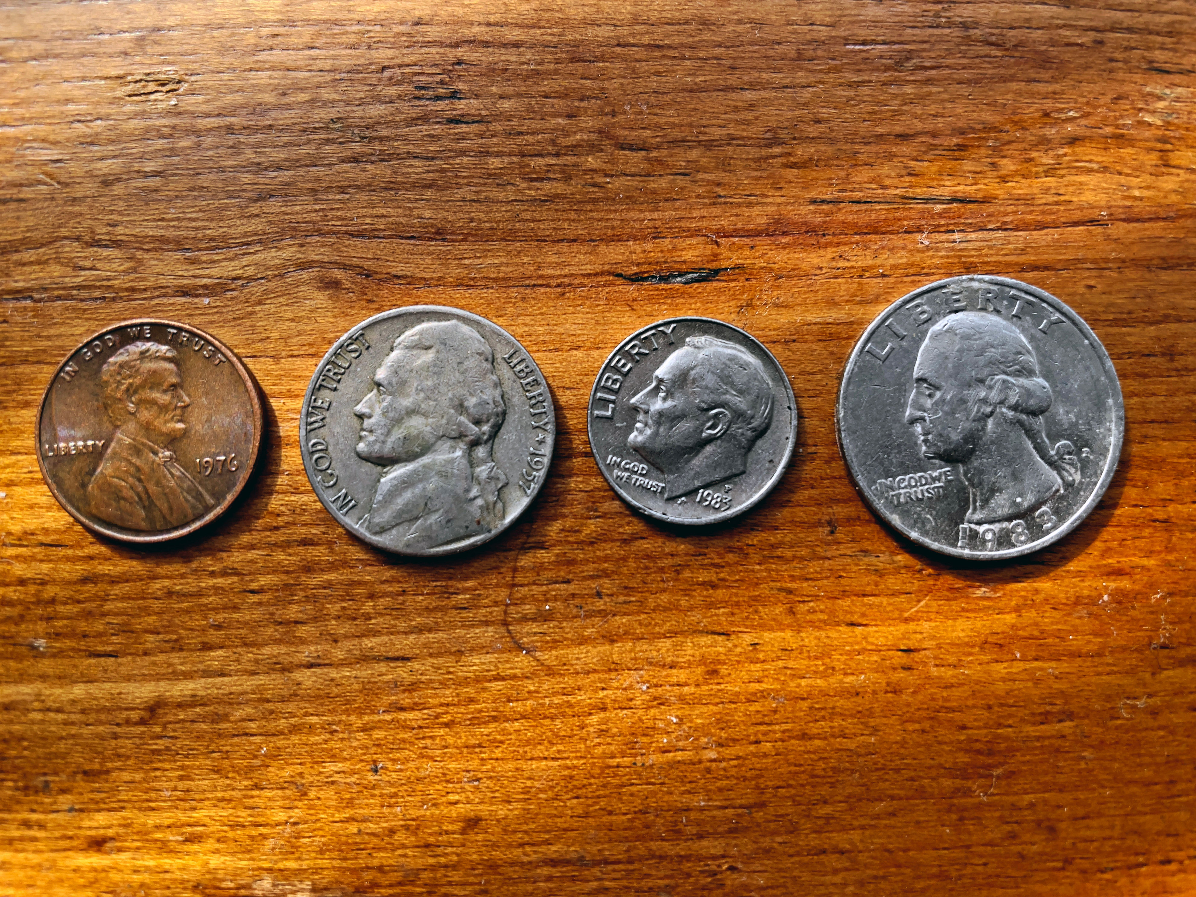 Four US coins are lined up on a wooden surface: a penny, nickel, dime, and quarter, each showing heads side with varying designs
