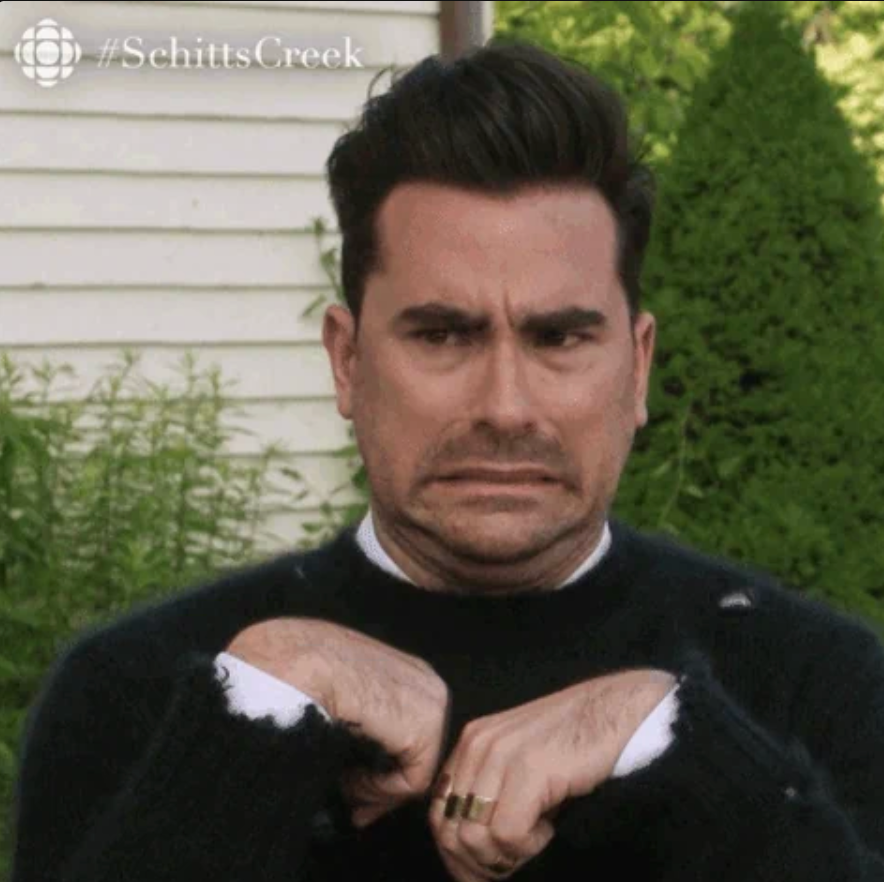 Man with an uncomfortable expression, holding hands awkwardly in front, dressed in a dark sweater. Background features greenery and a house wall