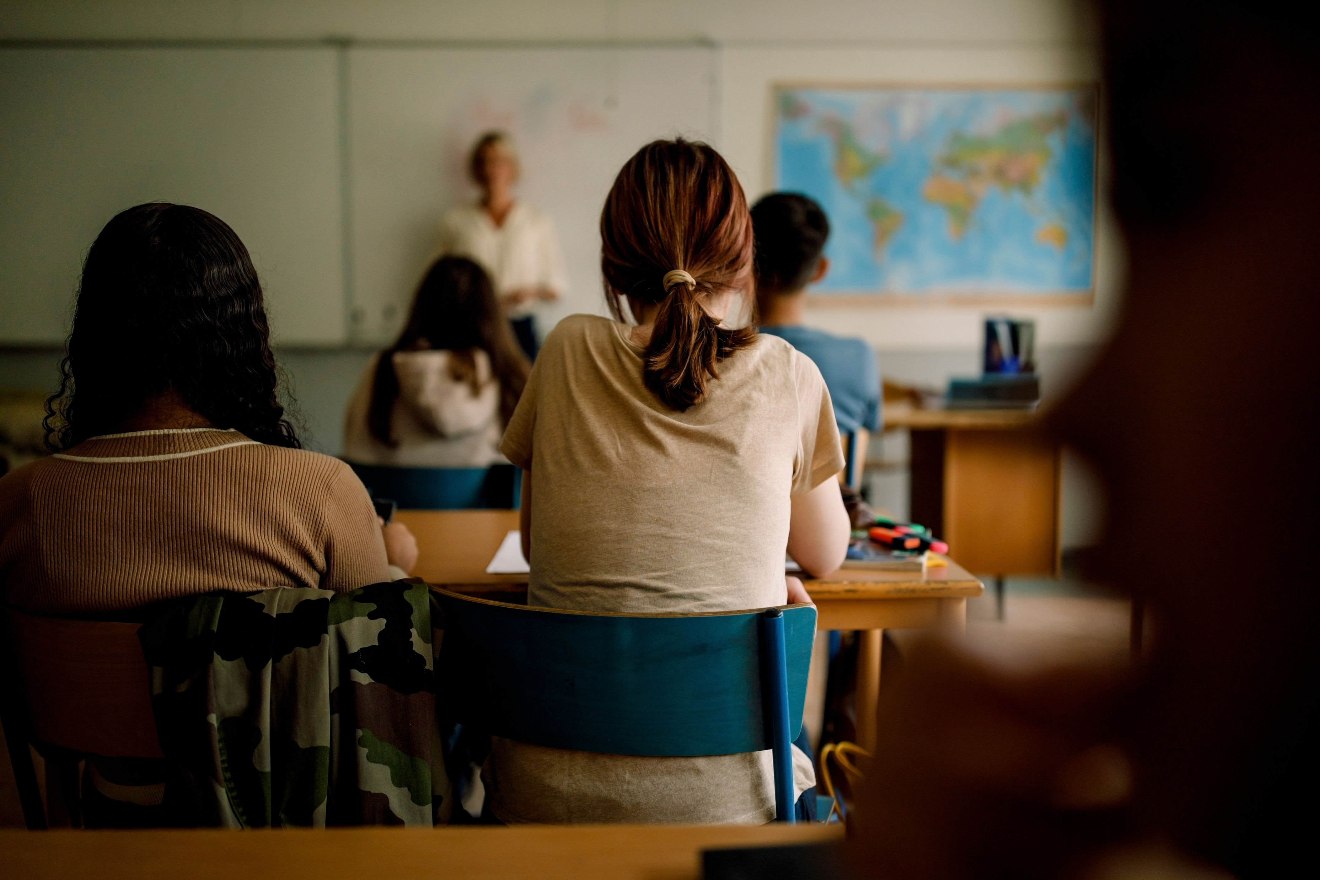 Students sit in a classroom, attentively facing a teacher at the front. A world map is on the wall