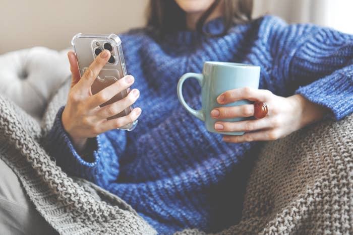 Person in cozy sweater holds a smartphone and mug, sitting on a couch wrapped in a knitted blanket