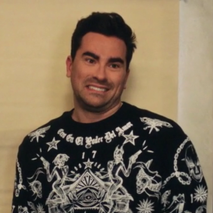 Person smiling awkwardly, wearing a graphic sweater with intricate patterns, from a scene in the show Schitt's Creek