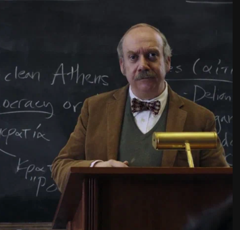 A man stands at a lectern in front of a blackboard with notes. He is wearing a tweed jacket, sweater, and bow tie, appearing as an educator or lecturer