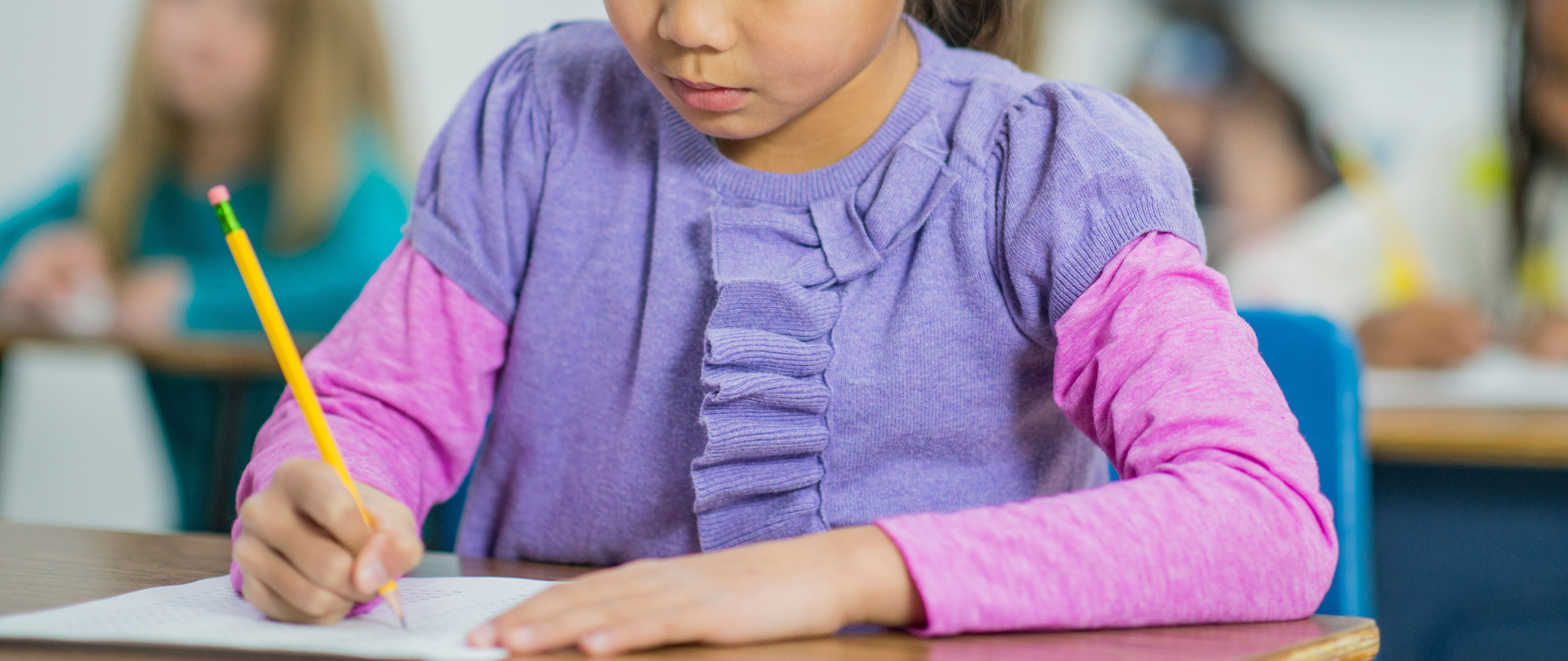 Young girl in a classroom focused on writing at her desk, with classmates blurred in the background