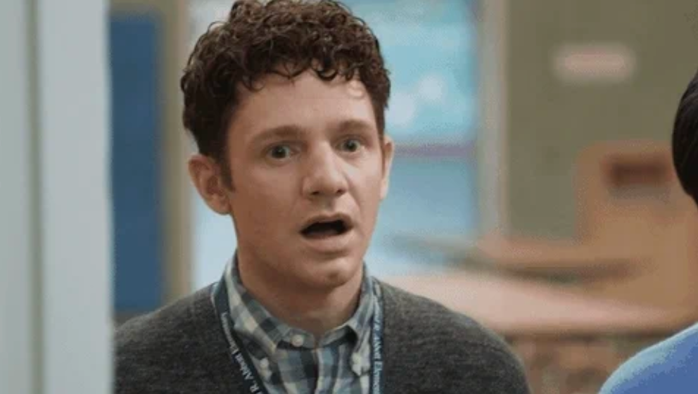 A surprised man with curly hair and a lanyard, wearing a checkered shirt and cardigan, stands indoors
