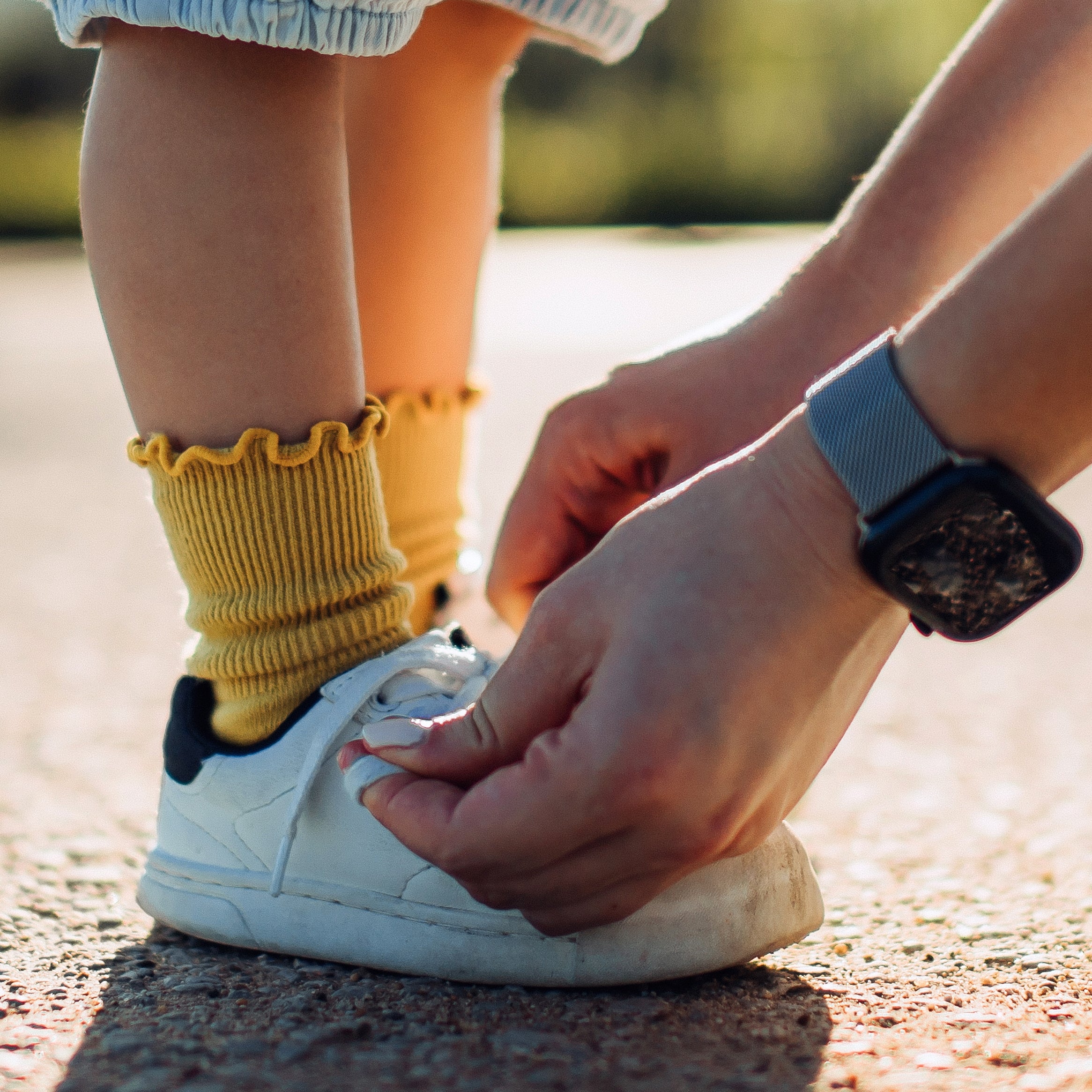 An adult is tying the shoelaces of a young child standing on a path. The child wears shorts and patterned socks