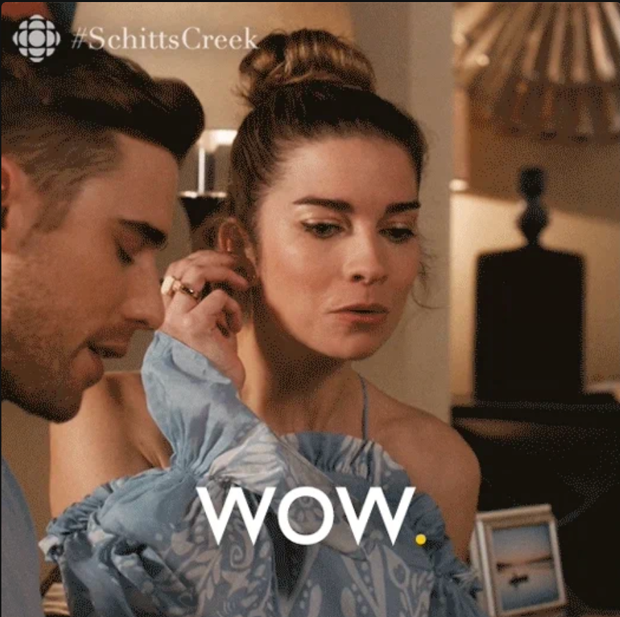 Two people from a TV show scene; one has short hair, the other has hair in a bun and wears a blue ruffled dress. “WOW” is written on the image