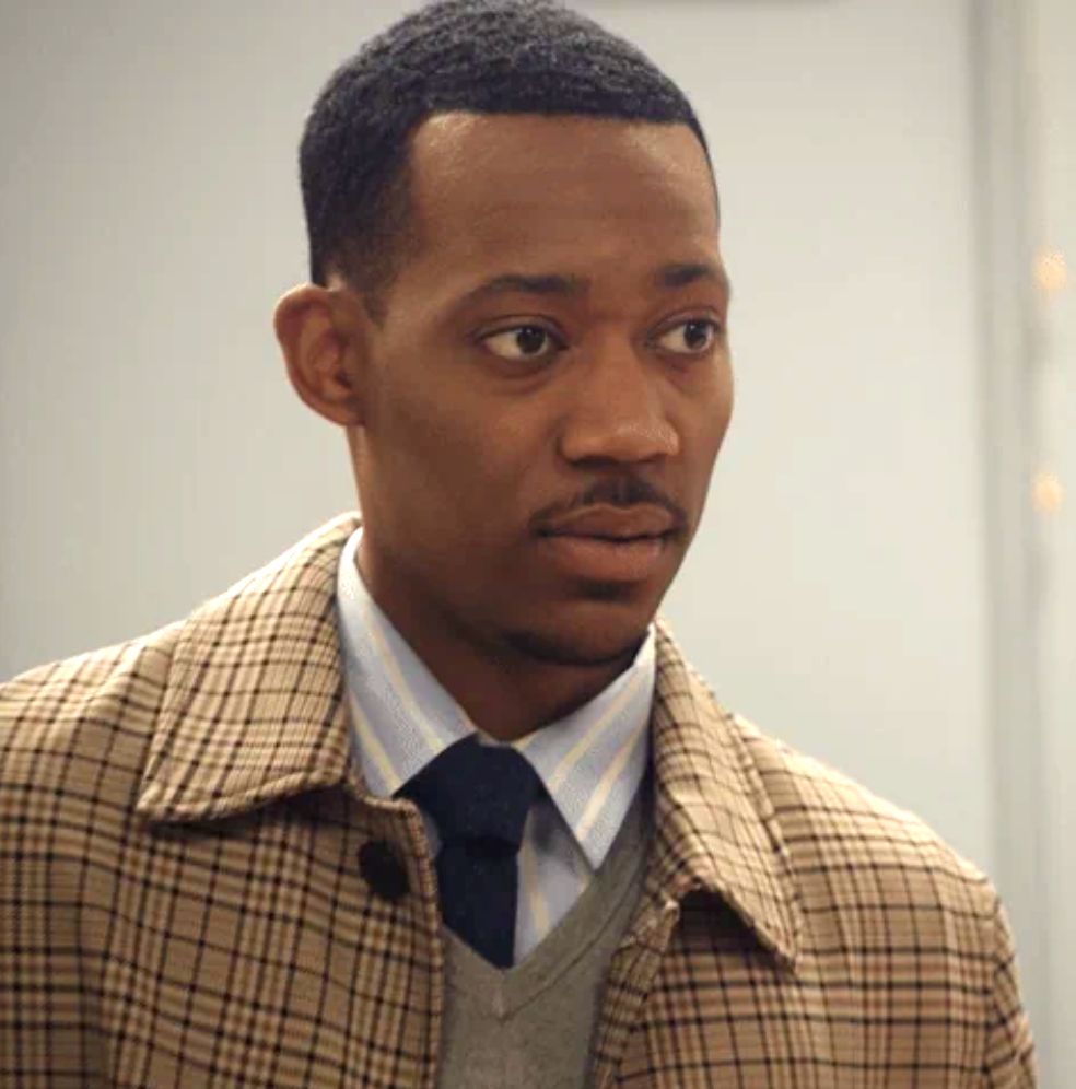 A person in a plaid coat and tie looks towards the camera against an indoor backdrop