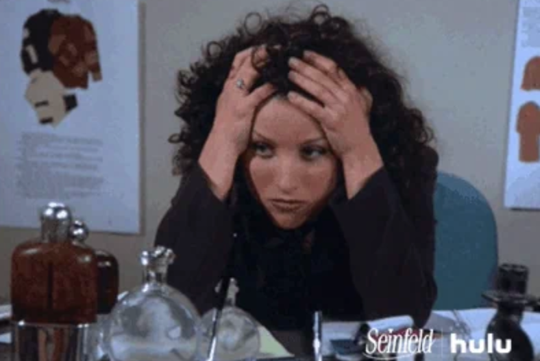 Person sitting at a desk, looking frustrated with their hands on their head, surrounded by various objects