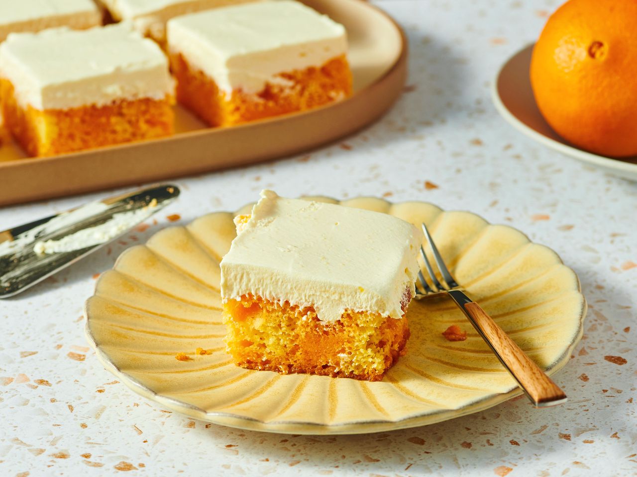 A piece of orange creamsicle sheet cake on a plate with a fork, other pieces and an orange in the background