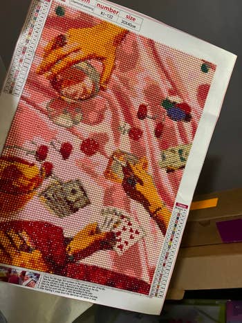 Hands holding playing cards on a pink background, surrounded by small containers, possibly for a diamond painting kit