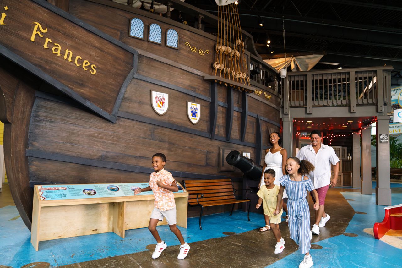 A group of children and adults walking near a large ship exhibit indoors