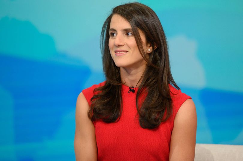 An individual with long, dark hair wearing a red sleeveless top is standing and smiling against a light blue background.