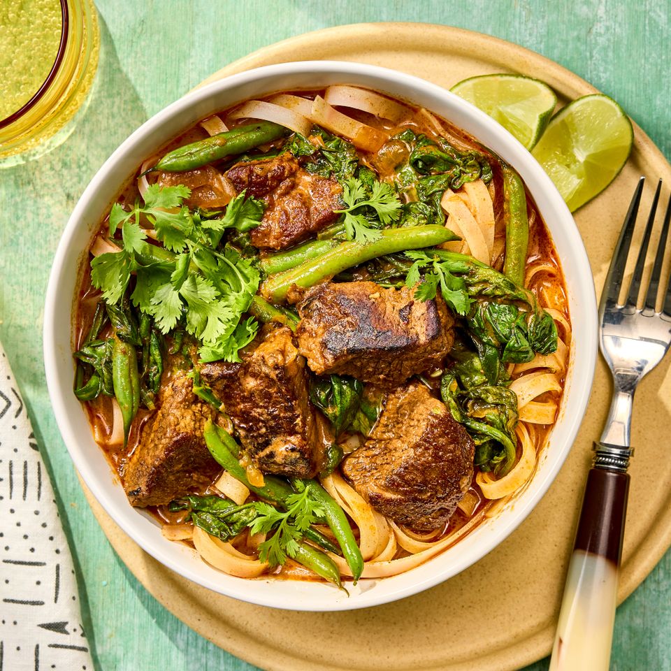 A bowl of beef curry with noodles and vegetables garnished with herbs served with lime wedges and a fork on the side