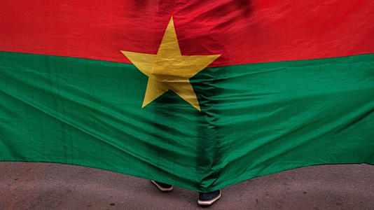 FILE - The Burkina Faso nationals flag with a man standing in front of it is seen during a memorial ceremony in Ouagadougou, Burkina Faso, Jan. 23, 2016.