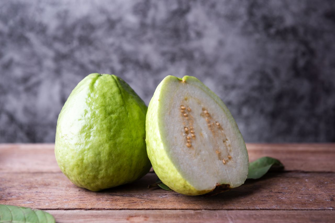 Whole guava fruit and a halved guava on a wooden surface with leaves nearby